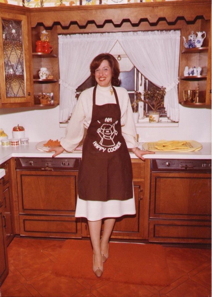 My paternal grandmother, Ruth Wildes, standing in her kitchen in an apron that reads, "I am a happy cooker."